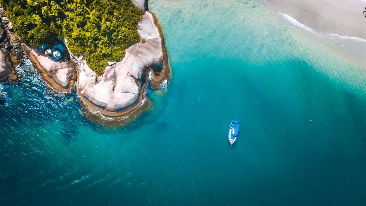 Vista de cima de uma praia com água azul turquesa, areia branca e formação rochosa com um barco na água. 