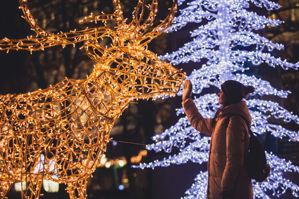 Una donna con cappotto e cappello accarezza una renna fatta di luci come parte della decorazione natalizia di una città