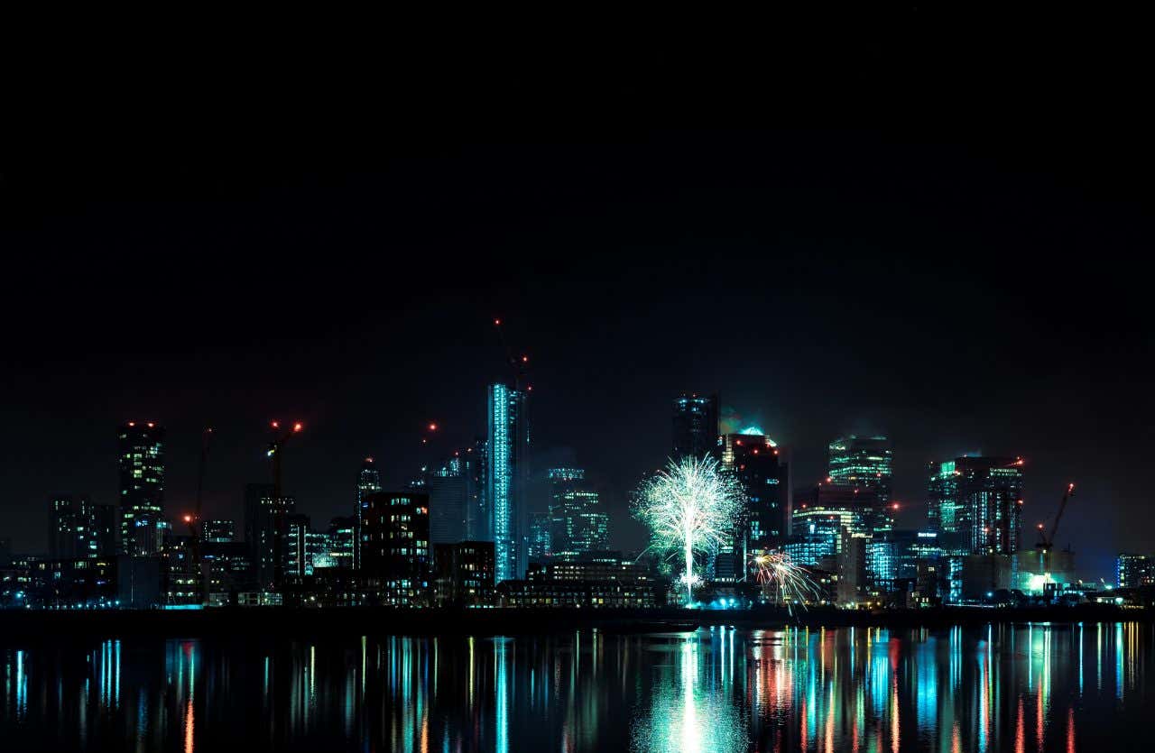 A view of London's high-rise skyline at night with fireworks reflecting in the river