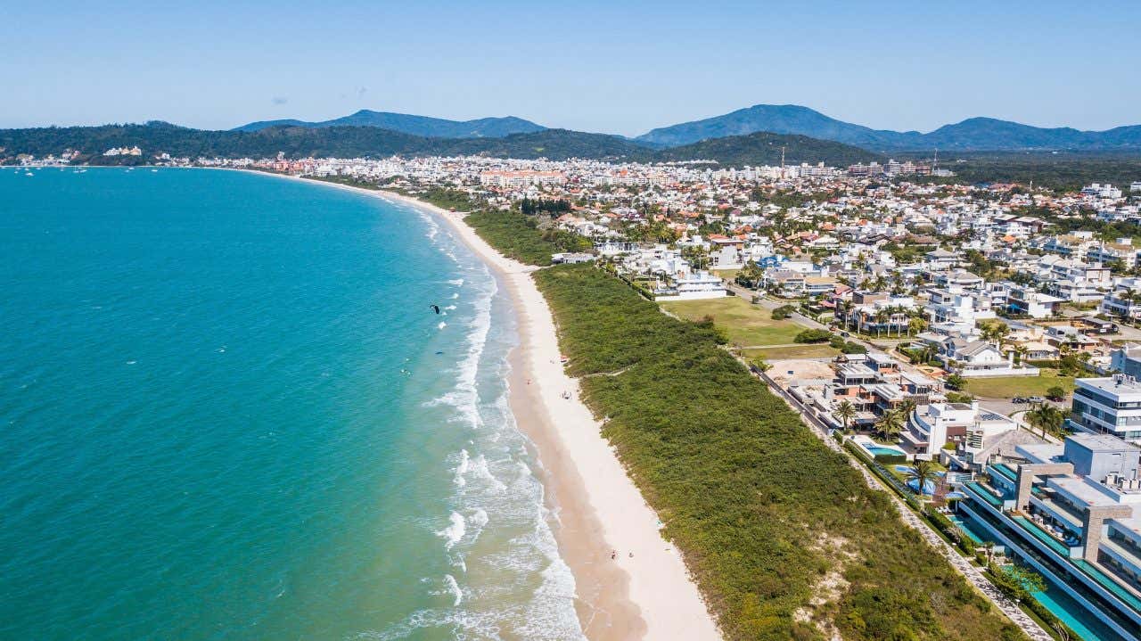 Vista aérea da praia Jurerê International com o oceano turquesa e areia branca à esquerda e as casas e hotéis luxuosos à direita