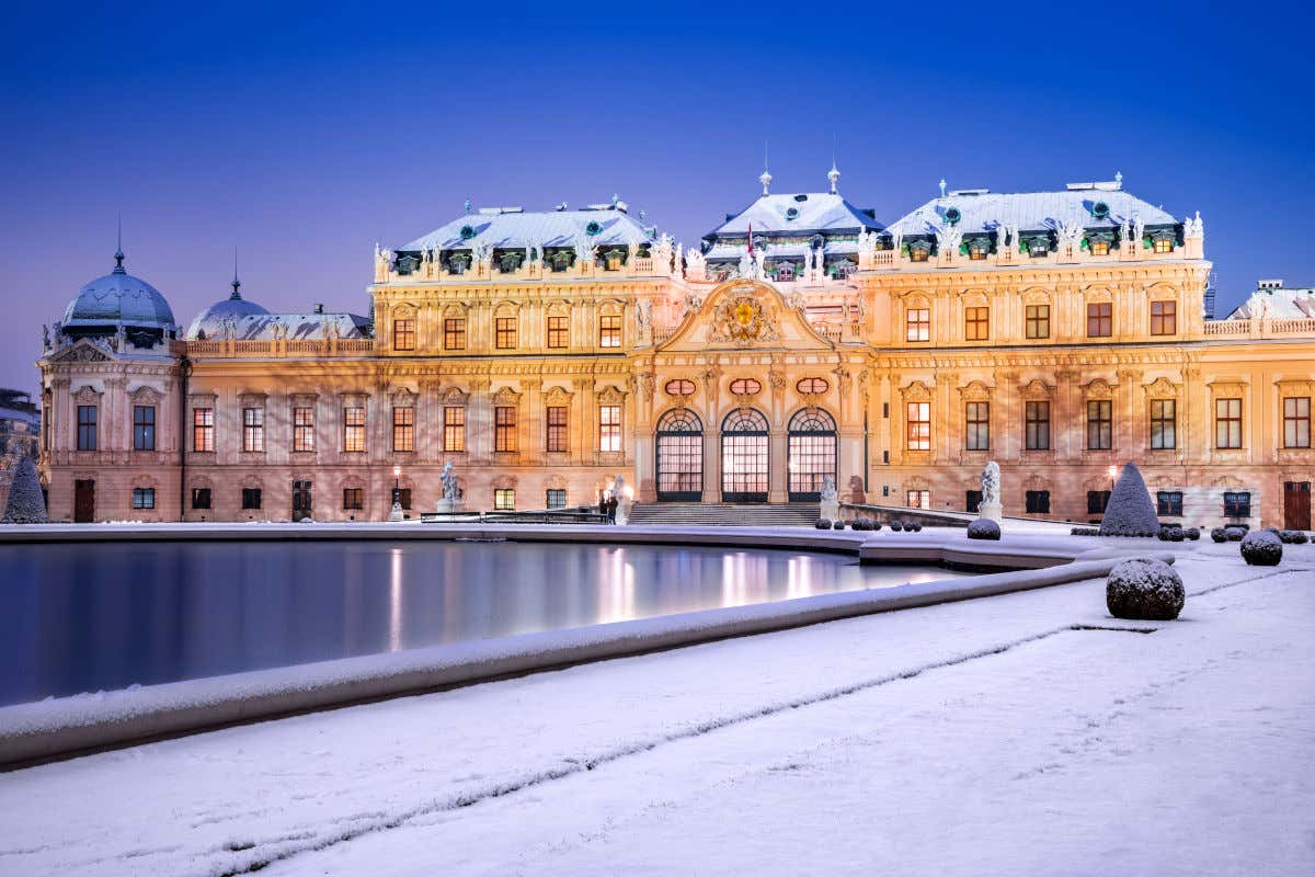 Exteriores de un palacio con sus tejados totalmente nevado y con un gran estanque rodeado de nieve por la noche