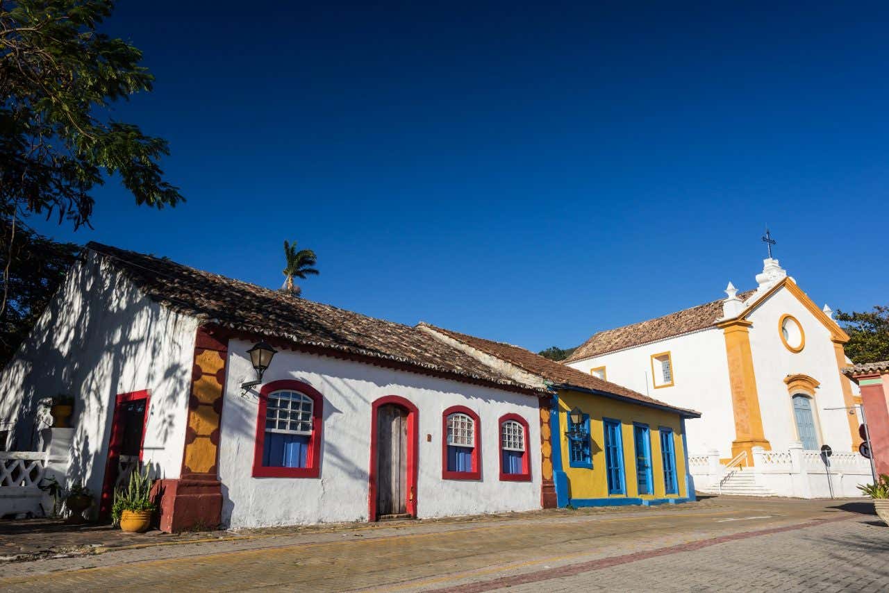 Duas casas coloniais coloridas e a igreja de Nossa Senhora das Necessidades em uma rua de paralelepípedos