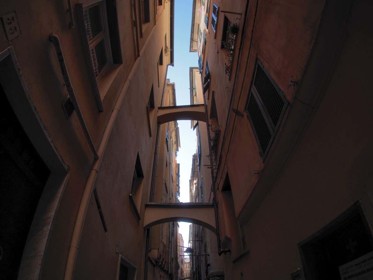 Vista dal basso di un vicolo molto stretto e buio del centro di Genova, composto da due palazzi di colore arancione molto vicini fra loro e collegati da due archetti posti all'altezza del primo e del secondo piano