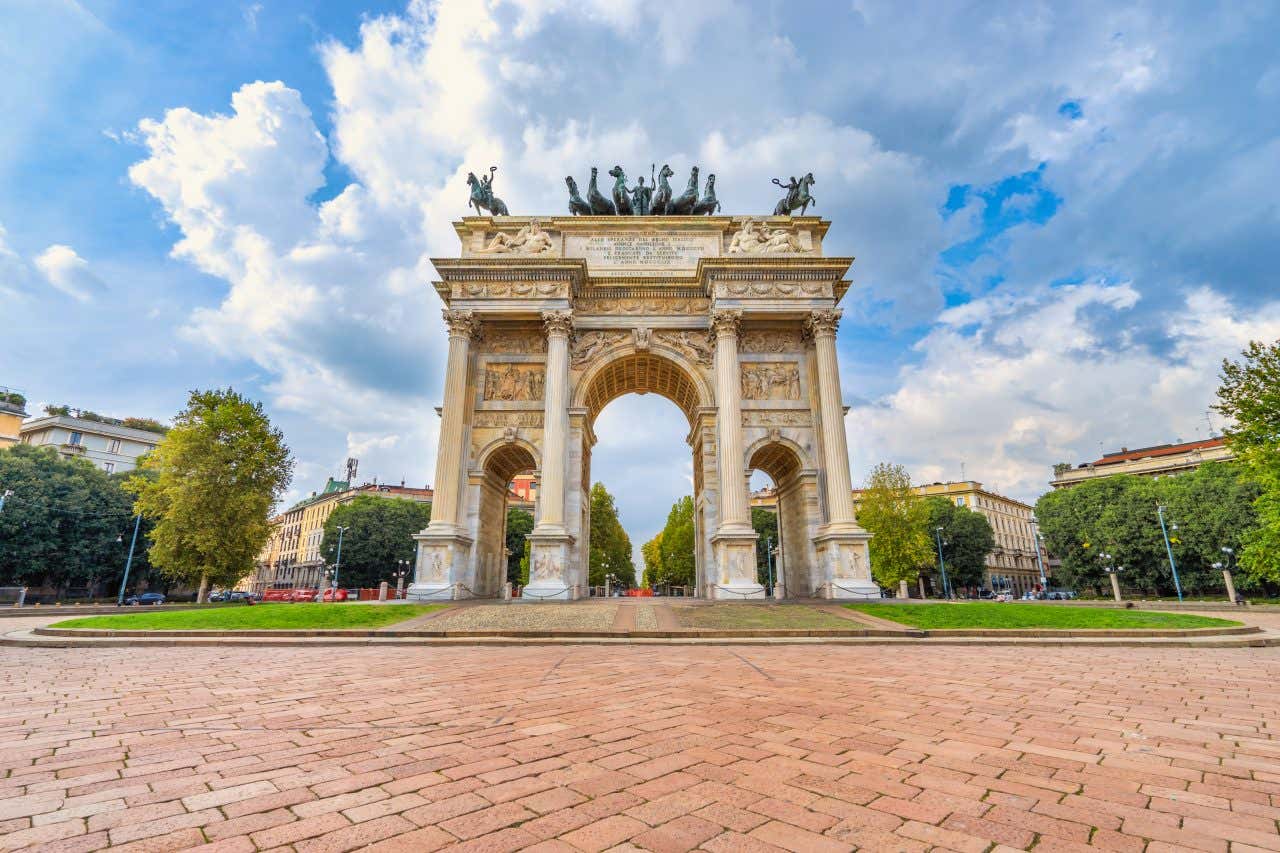 Facade of a large marble arch with bronze sculptures on top on a sunny day with buildings in the background