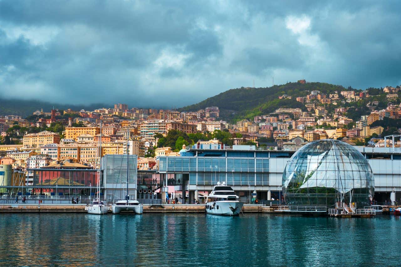 Un porto con uno yatch armeggiato davanti a un molo con diversi edifici dall'aspetto futuristico. Uno sulla destra è a forma di sfera ricoperta di vetrate trasparenti. Subito dopo si vede l'edificio che ospita l'Acquario di Genova. Sullo sfondo la città che si arrampica sulle verdi colline in una giornata nuvolosa