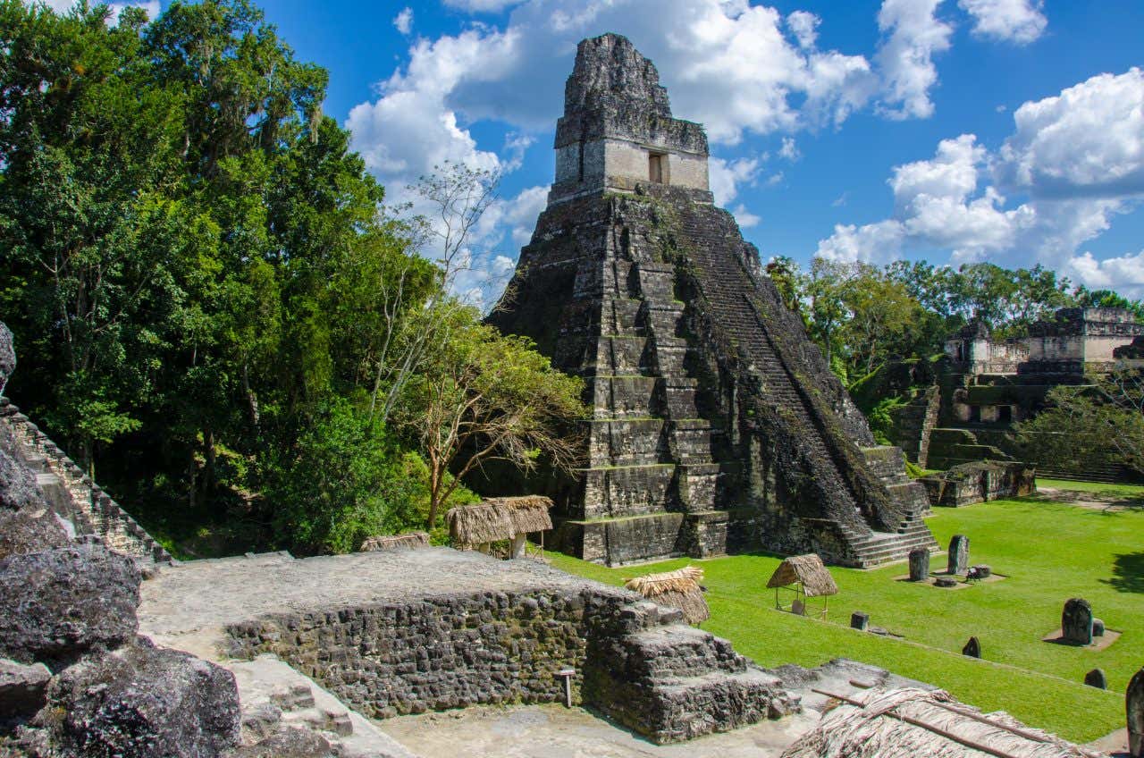 A tall Maya temple in Tikal with some smaller structures in the foreground and a cloudy sky in the background.