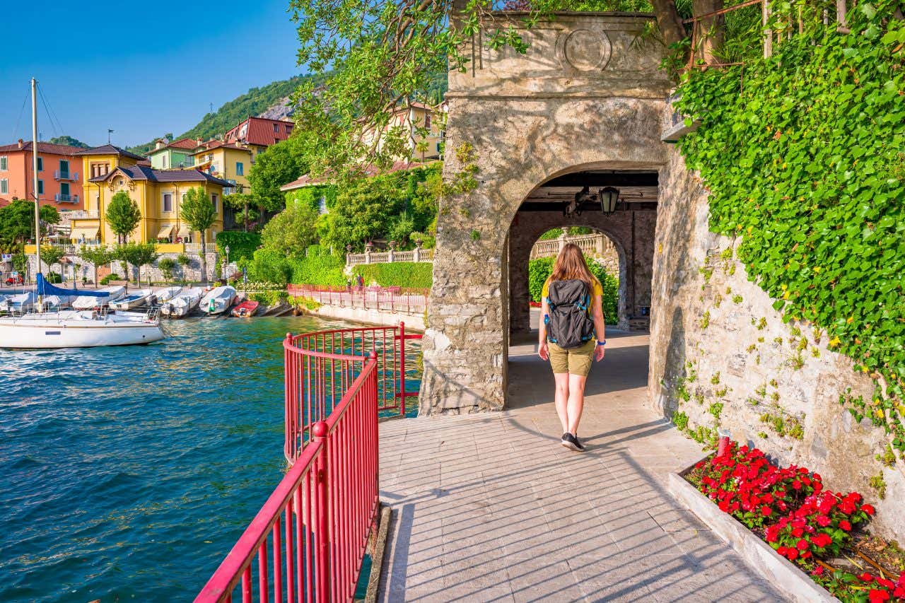 A woman with her back turned walking on a wooden path next to a lake with boats and colorful buildings on the left 