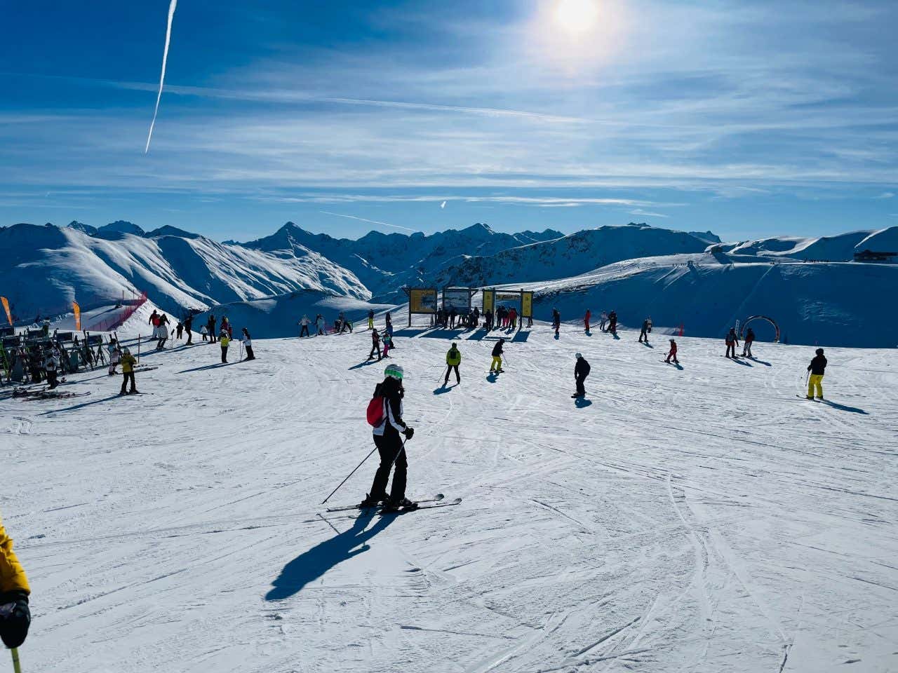 Un altopiano innevato con diverse persone che si spostano sugli sci e, sullo sfondo, montagne bianche e il sole nel cielo limpido