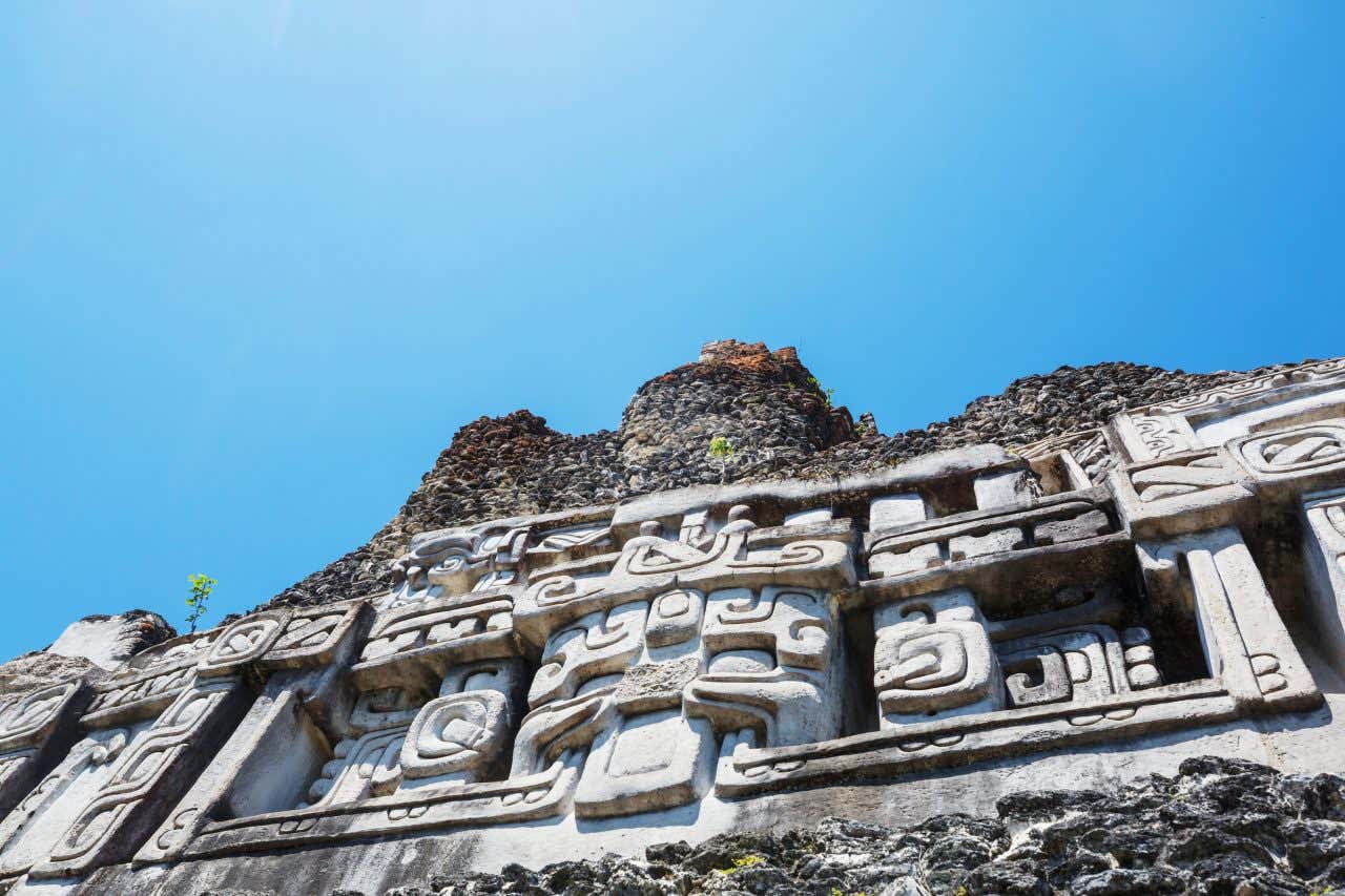 Maya ruins in Xunantunich as seen from below, with an ornate face carved into a wall and a clear blue sky above.