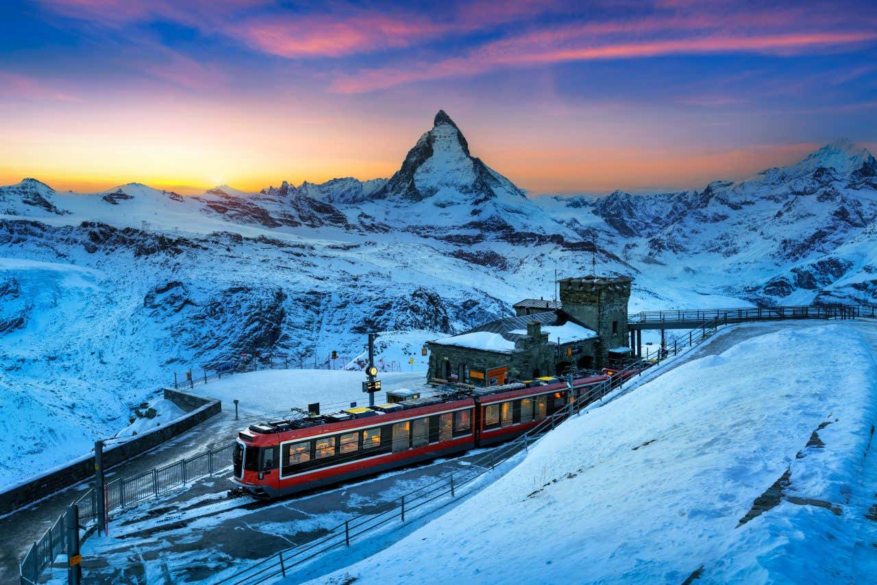 Una cima innevata leggermente incurvata di un monte che si staglia sul tramonto multicolore e, in primo piano, un piccolo treno rosso fermo a una stazione in pietra