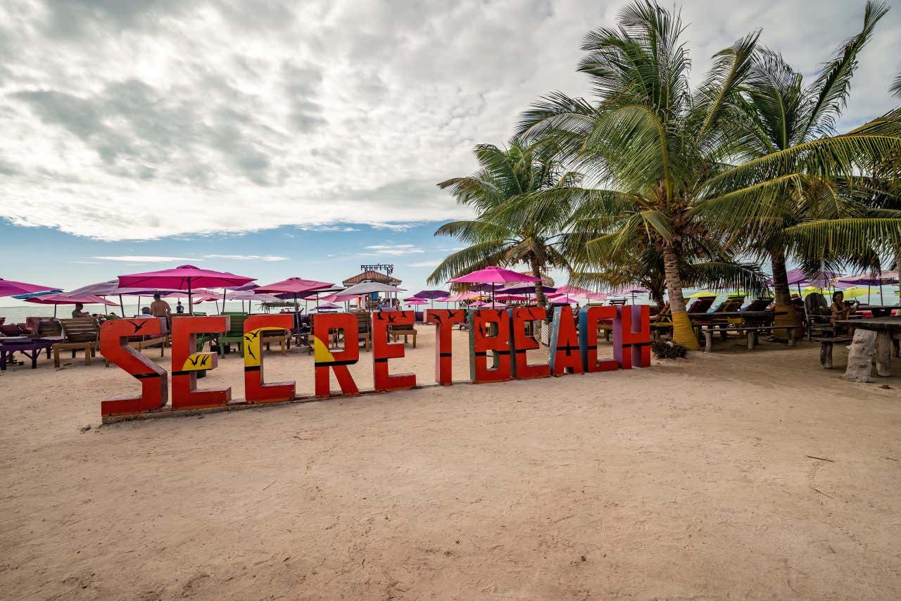 The sign for Secret Beach on Secret Beach with some palm trees on the righthand side and a cloudy sky in the background.