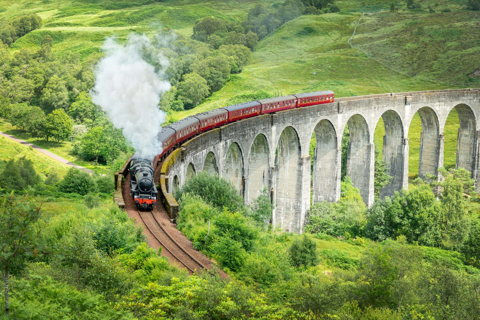 Una locomotiva di colore rosso percorre un ponte di pietra nella vegetazione scozzese