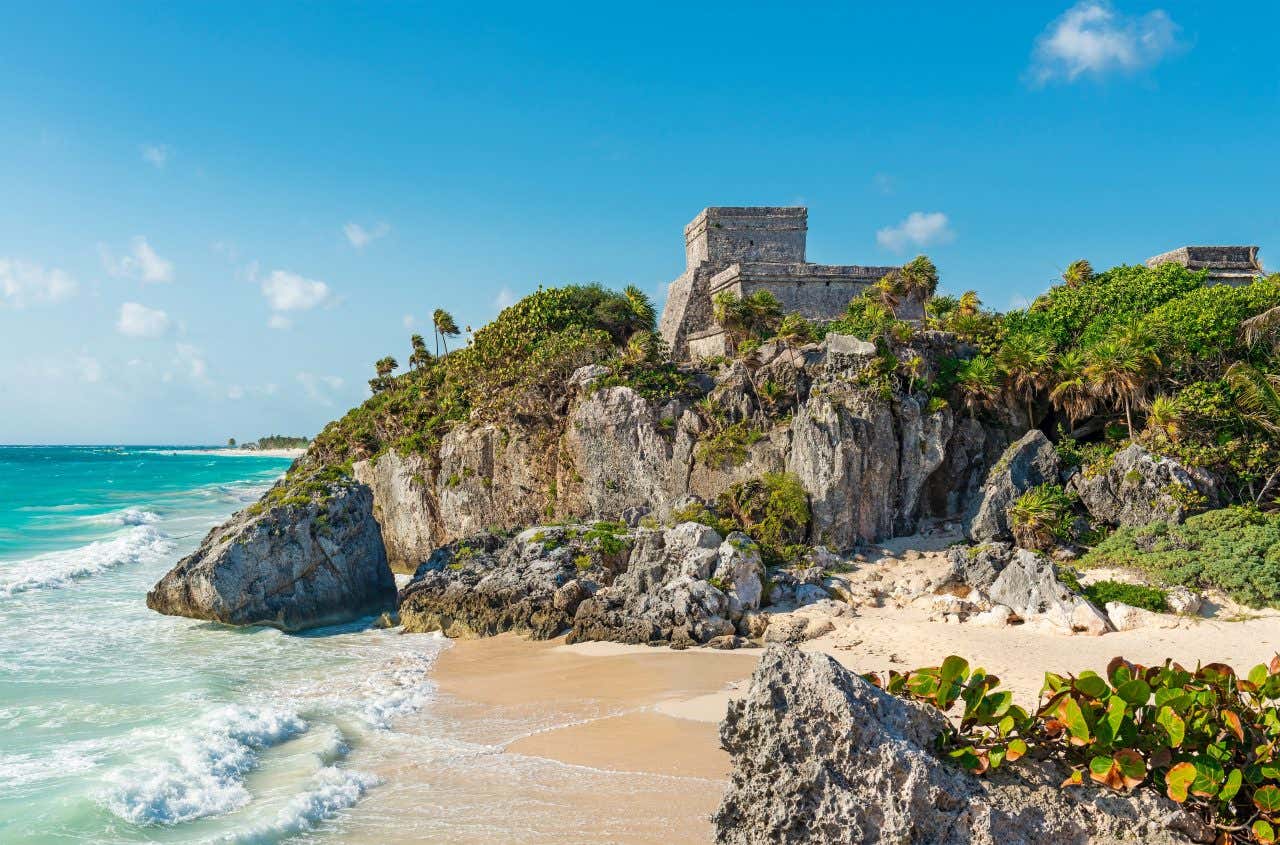 As ruínas de Tulum na beira do penhasco, com um céu azul levemente nublado ao fundo