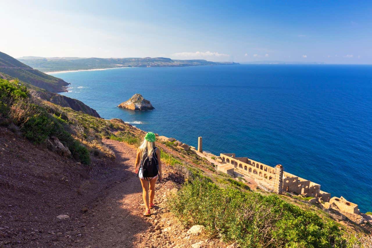 Una ragazza di spalle cammina lungo un sentiero costiero per raggiungere i resti di una fabbricato affacciato sul mare in un giorno di sole