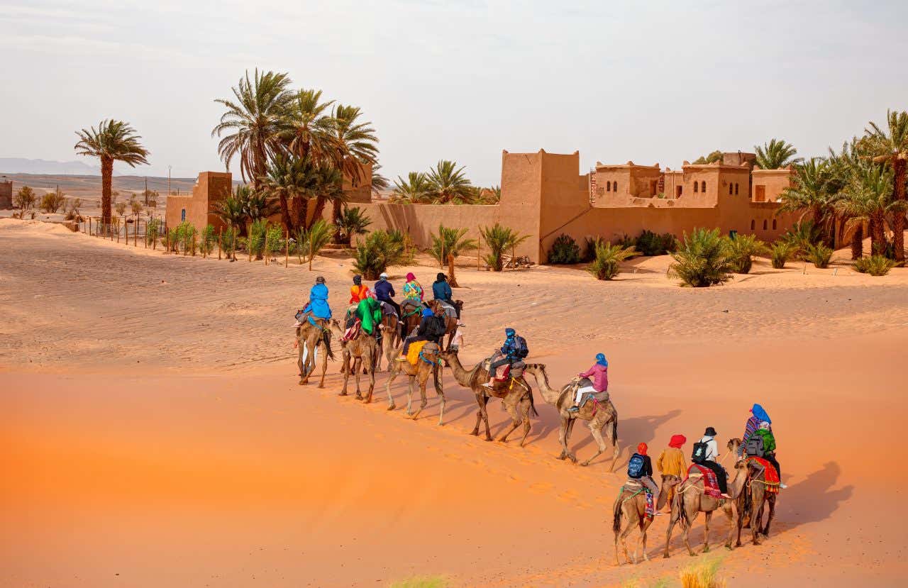 Una carovana di persone in sella a dei cammelli attraversa il deserto marocchino e passa accanto a un piccolo insediamento circondato da palme