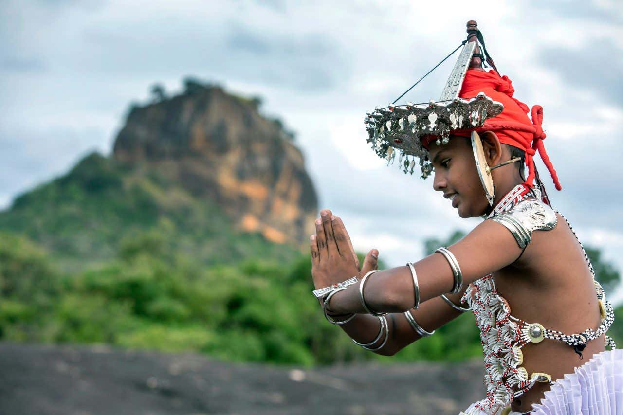 Un bambino srilankese in abiti tradizionali ripreso di profilo mentre giunge le mani durante l'esecuzione di una danza tradizionale. Sullo sfondo di intravede un monte in una giornata parzialmente nuvolosa