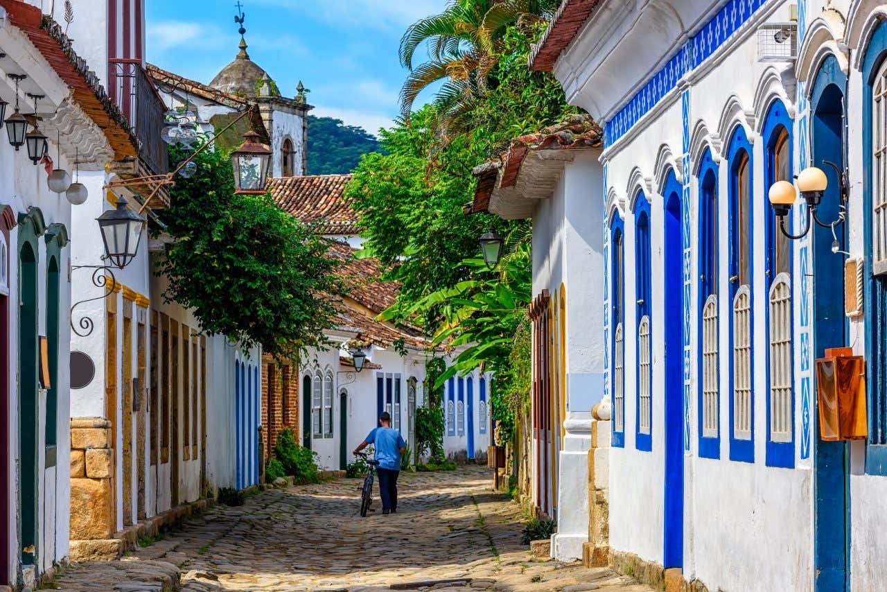 Um homem andando do lado de uma bicicleta em uma das ruas pé de moleque de Paraty, cercado de edifícios em estilo colonial