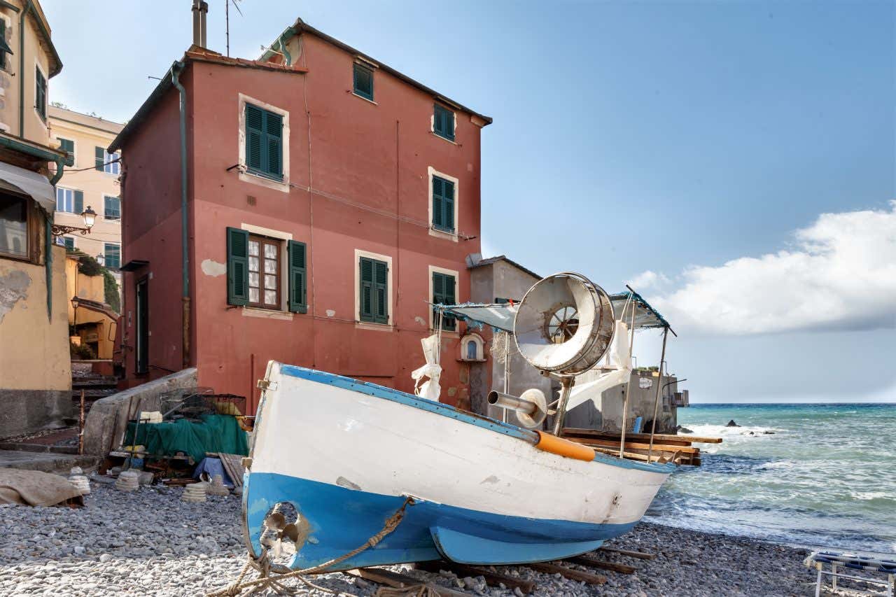 Um barco de pesca de madeira azul e branco abandonado em uma praia de seixos. Ao fundo, o mar levemente agitado e uma pequena casa vermelha com janelas verdes