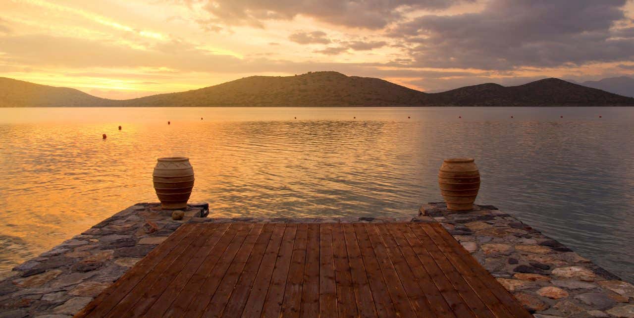 Una pedana in legno e pietra si interrompe per affacciarsi sulla Baia di Mirabello, a Creta. Nel cielo si percepisce un tramonto leggermente nuvoloso