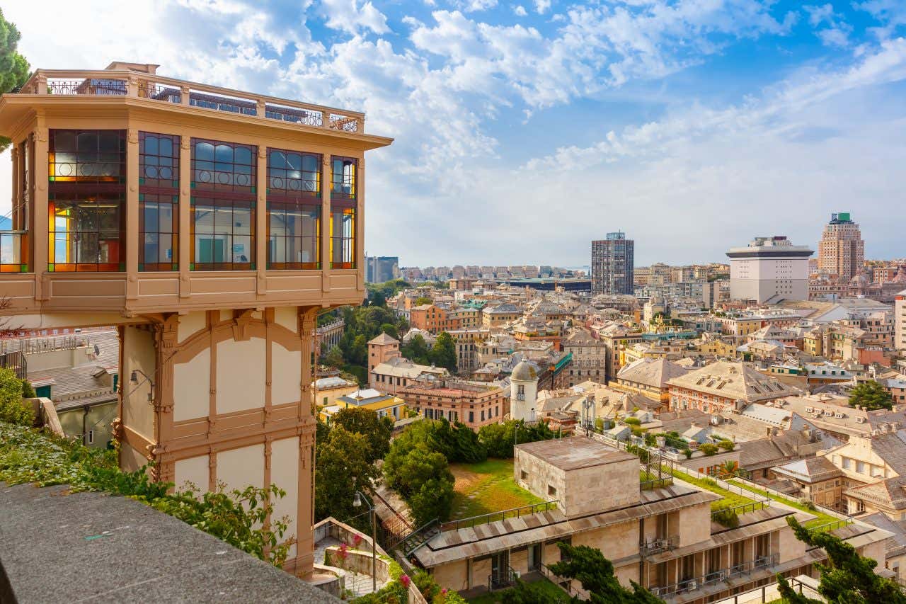 A cidade de Gênova fotografada de cima em um dia ensolarado. Os telhados e alguns arranha-céus podem ser vistos no horizonte. Em primeiro plano, é possível ver um pedaço de parede e um elevador Art Nouveau