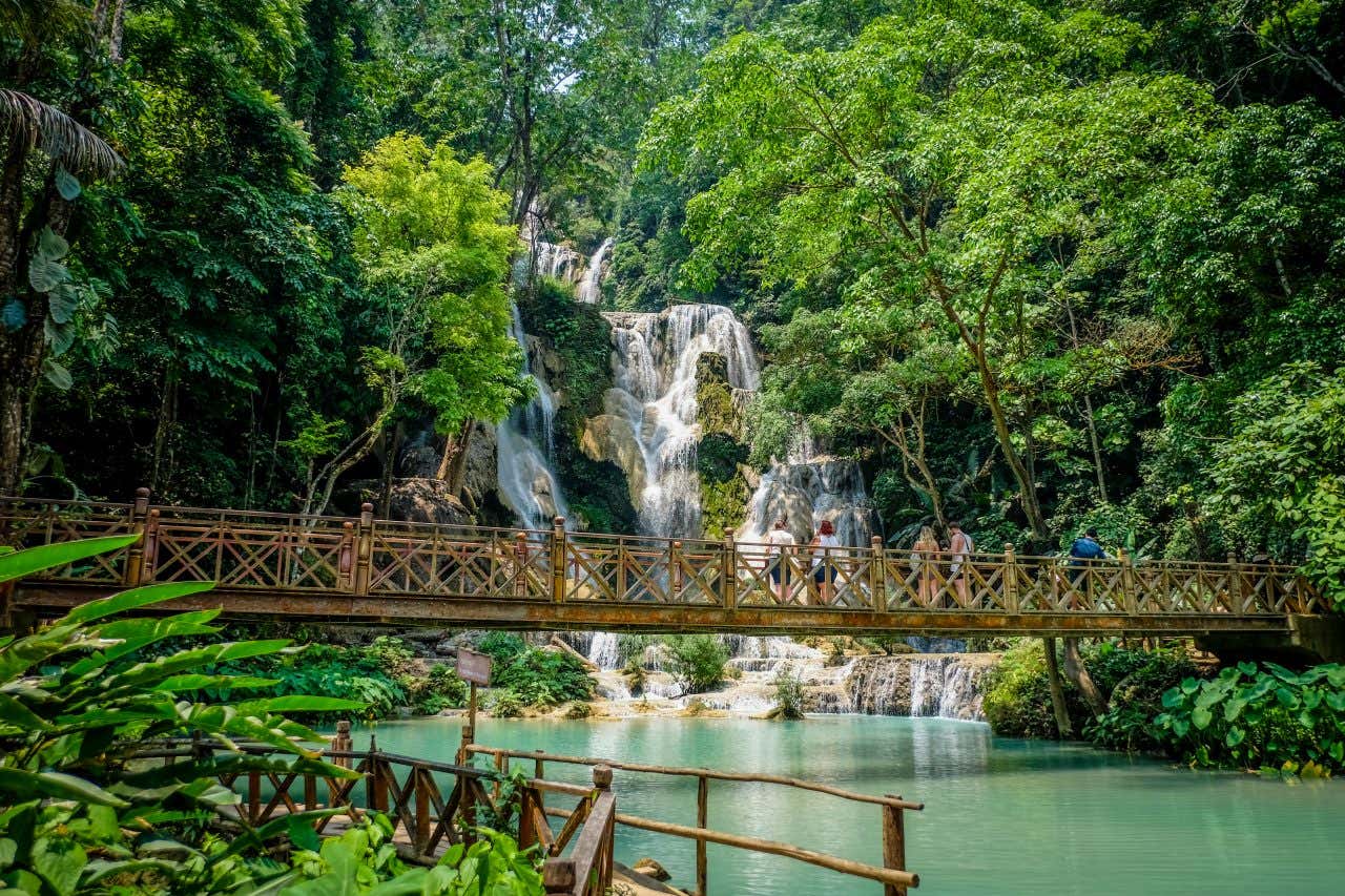 Un ponte in ferro attraversato da alcuni turisti che osservano i getti di una cascata all'interno di una fitta giungla in un giorno di sole