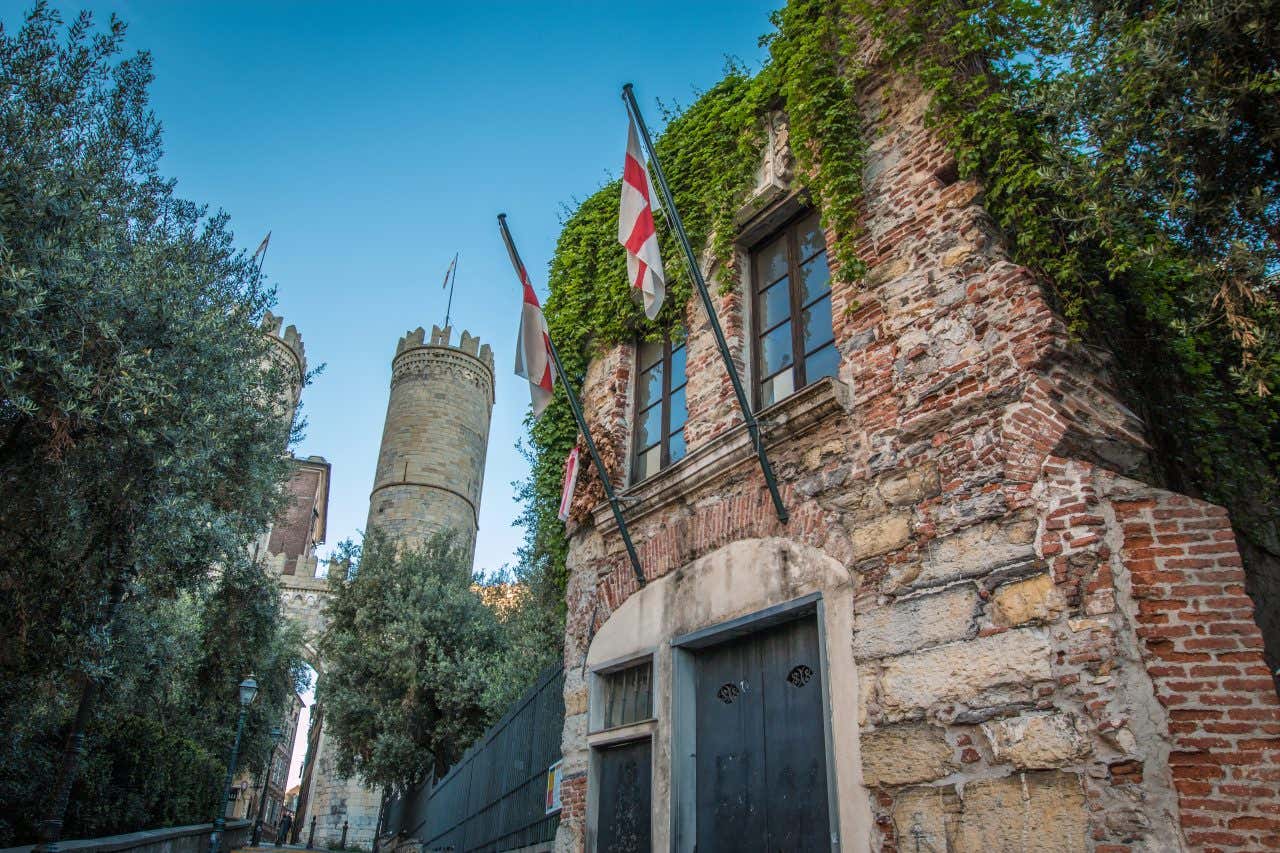 Fachada de tijolos de uma casa antiga coberta por hera e com dois mastros com uma cruz vermelha em um fundo branco. Atrás dela, há duas torres brancas com ameias que se destacam de uma oliveira em primeiro plano