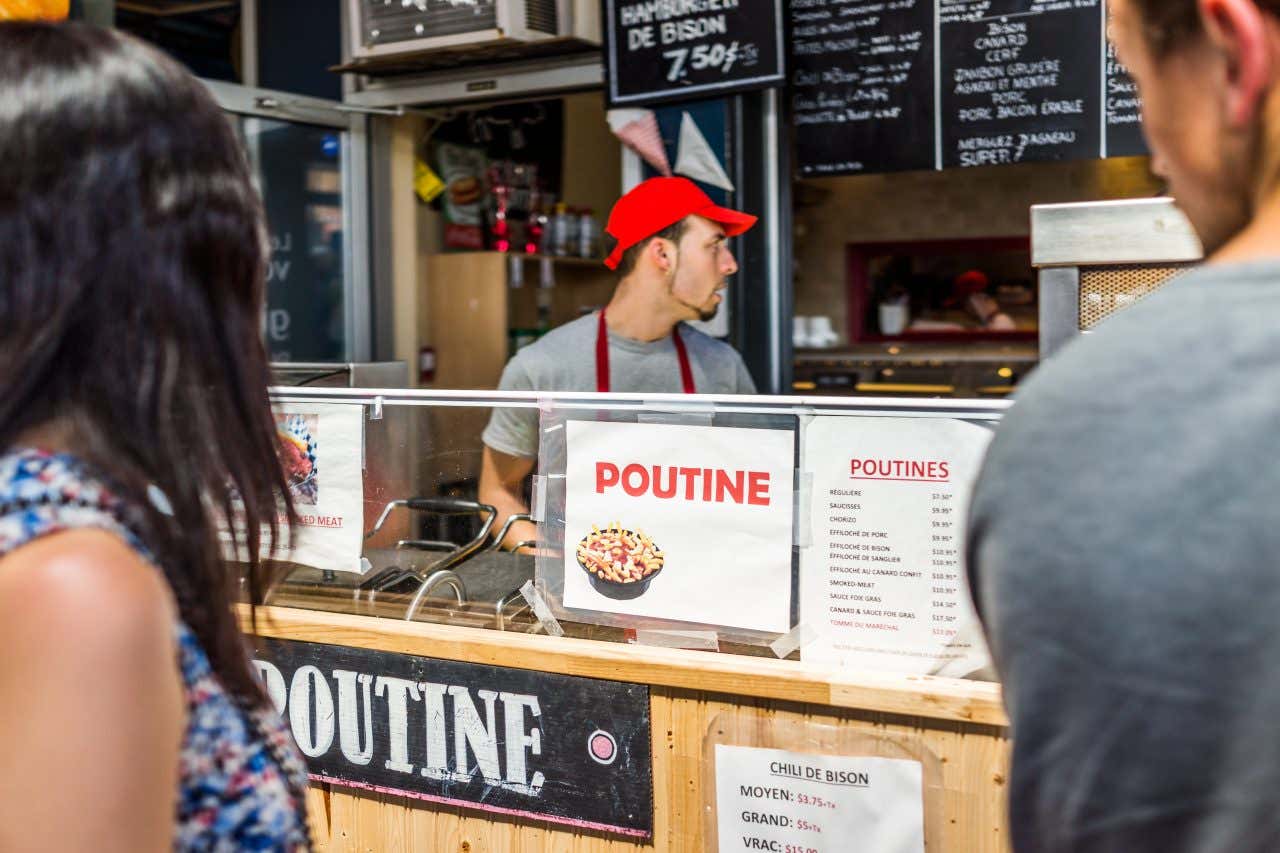 Un chiosco di poutine a Montréal, con due clienti in primo piano e un cameriere con un berretto rosso dietro il bancone.