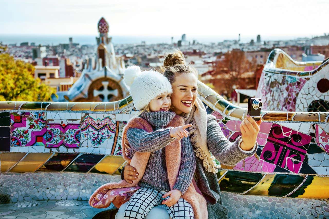 Una mamma tiene in braccio una figlia, entrambe in abiti invernali, sedute su una panchina decorata con piastrelle colorate. Stanno scattando una foto con la macchinetta fotografica e sul fondo si intravedono le cime dei palazzi del centro di Barcellona