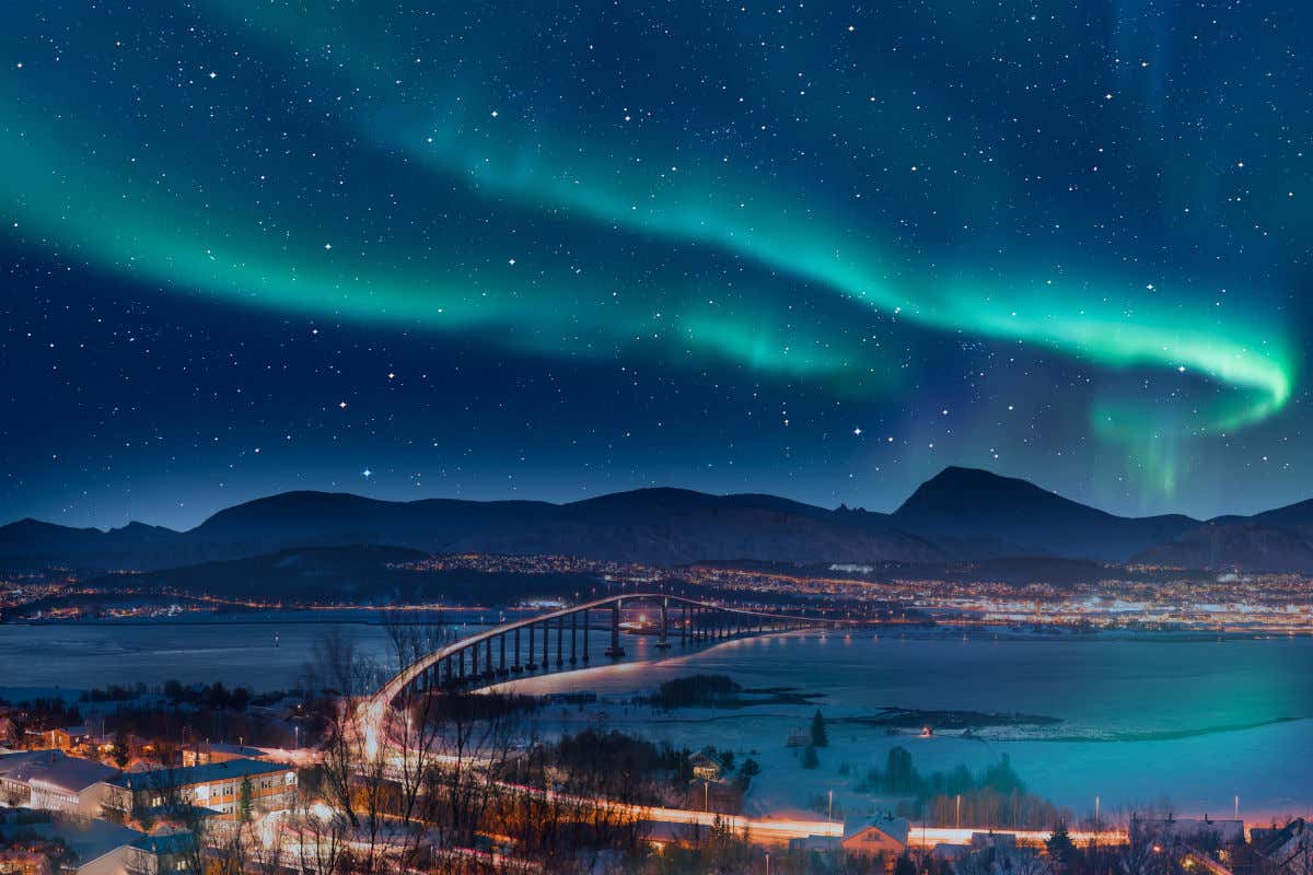 Aurora boreal y estrellas iluminando el cielo nocturno de la ciudad de Tromsø, con varias casas y un puente sobre el mar