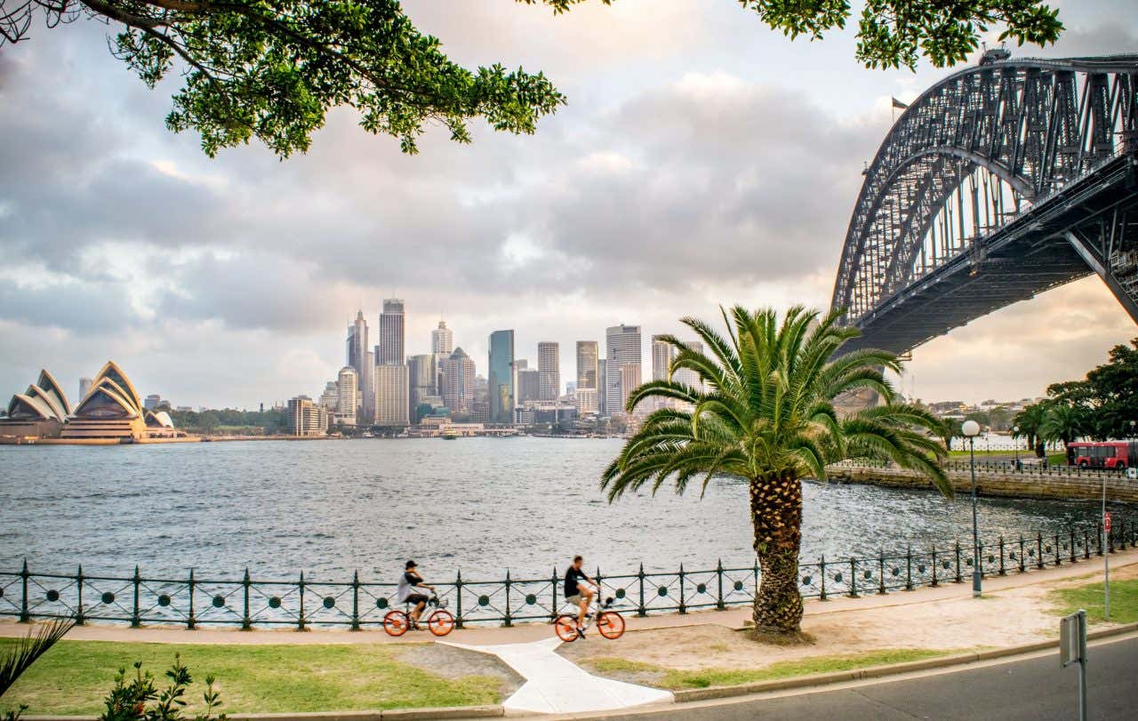 Duas pessoas andando de bicicleta por um passeio marítimo perto da Harbour Bridge com a Opera House e a cidade ao fundo