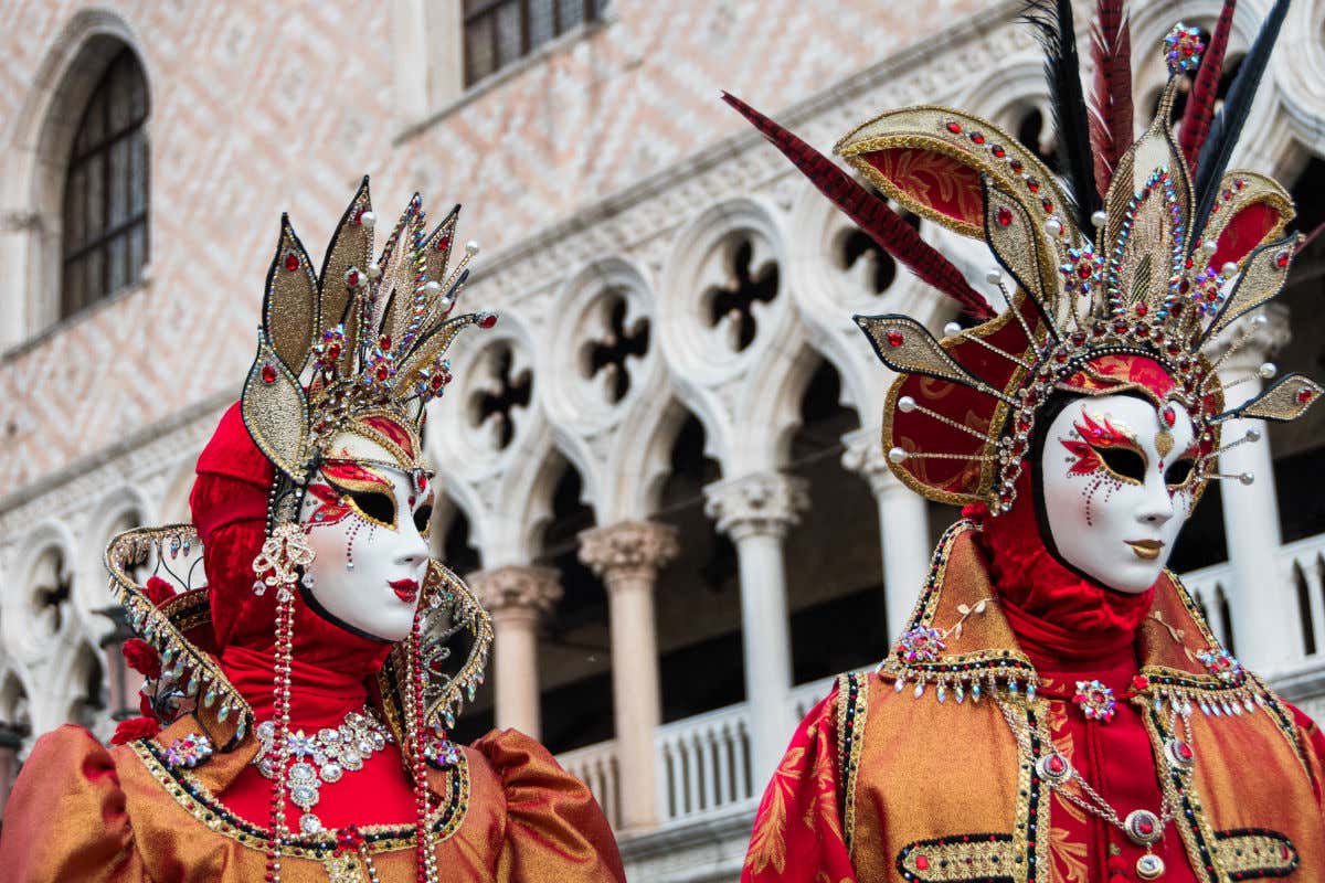 Two people dressed in costumes and typical Venetian masks in front of a building full of columns.