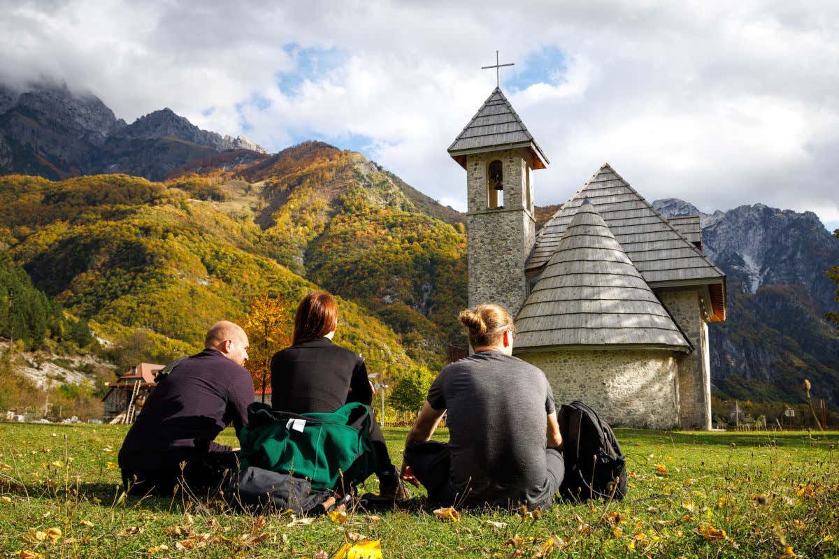 Tres turistas sentados en el césped contemplando una pequeña iglesia rodeada por montañas