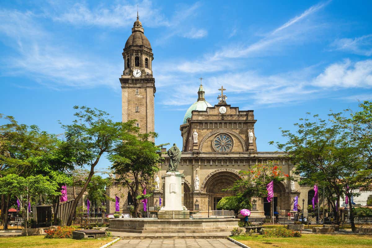 Panorámica de la catedral de Manila, con su rosetón, su cúpula y su torre en una plaza arbolada