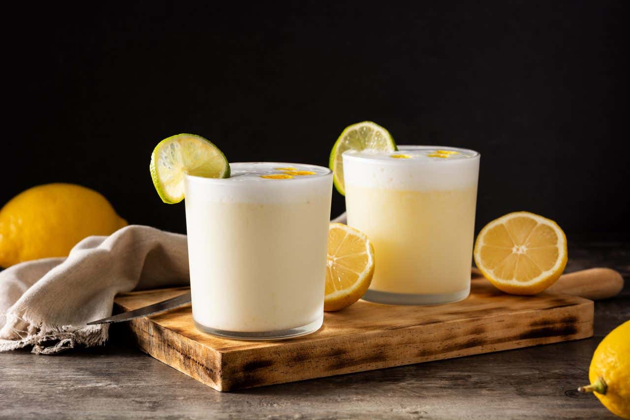 Dois copos com pisco sour sobre uma tábua de madeira e decoração de limões amarelos.