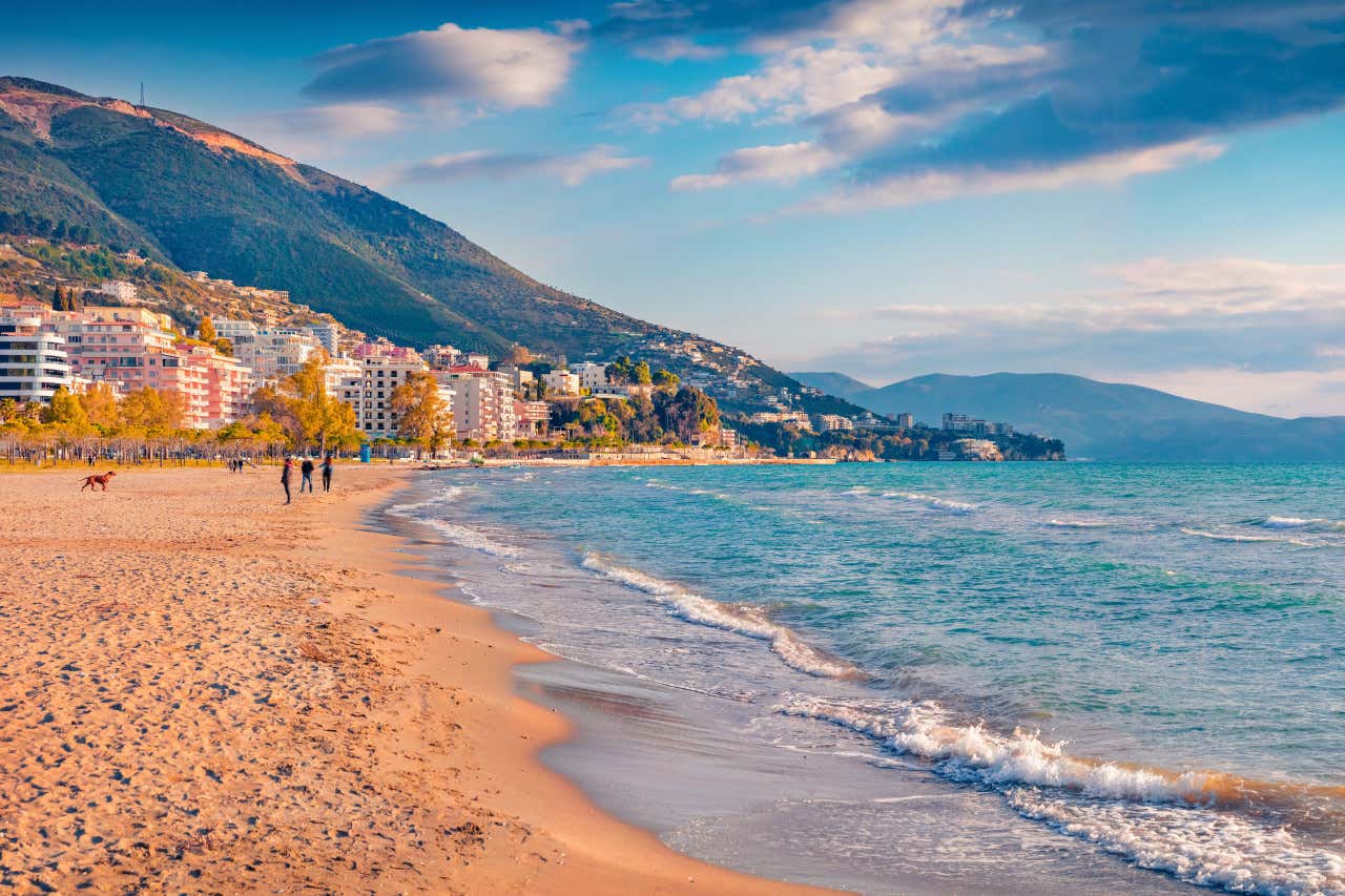 La spiaggia di Valona, di sabbia fine e dorata e mare calmo, con diversi edifici sul lungomare e montagne sullo sfondo