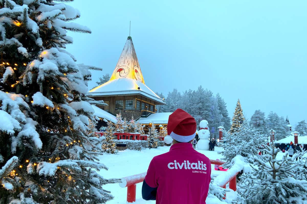 Un joven con un polo rosa con las letras de Civitatis y un gorro de Papá Noel en un paisaje repleto de nieve con árboles de Navidad y algunas cabañas