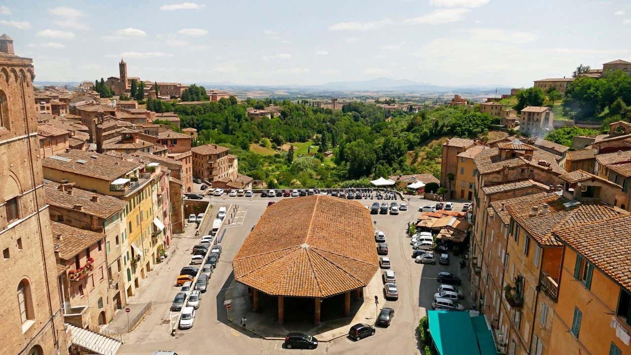 Una piazza con al centro una tettoia ovale e sullo sfondo verdi colline e il monte Amiata in lontananza coperto da una leggere foschia