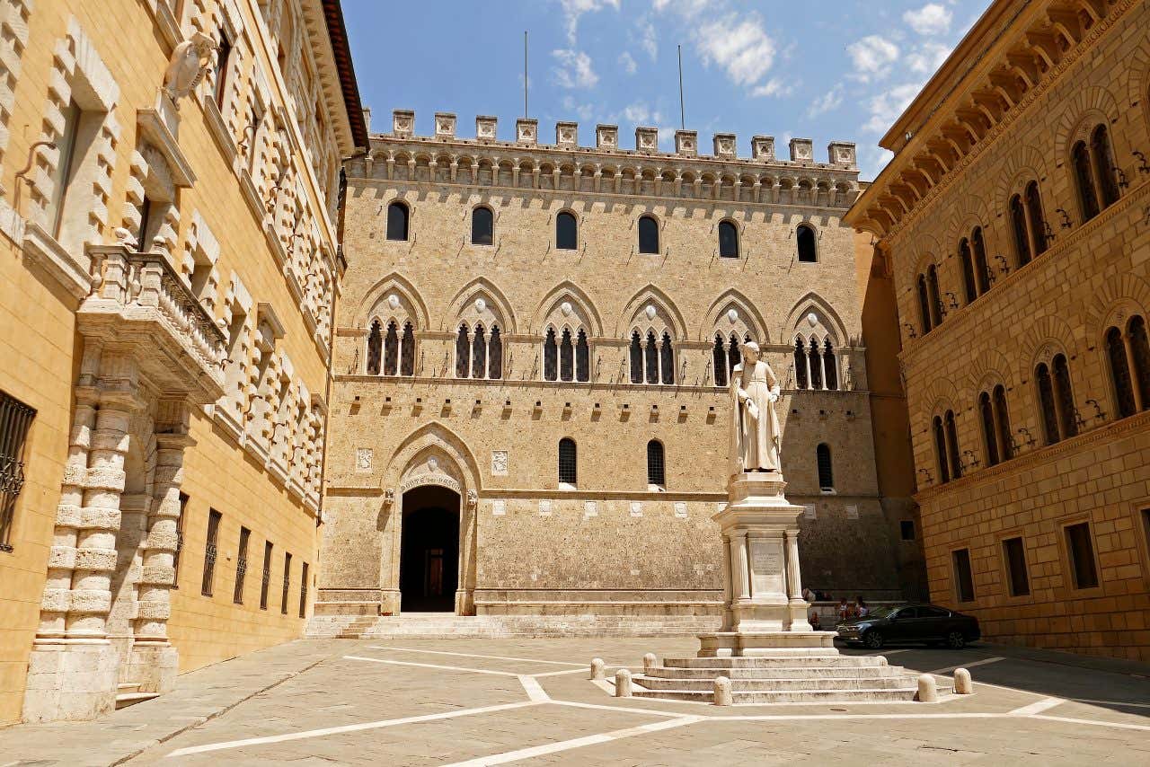 Un palazzo in stile gotico incastonato tra due palazzi in una piazza del centro di Siena in una giornata di sole, al centro della piazza la statua di un uomo.