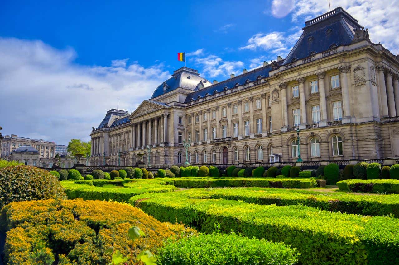 Fachada do Palácio Real sob um céu azul com os jardins em primeiro plano