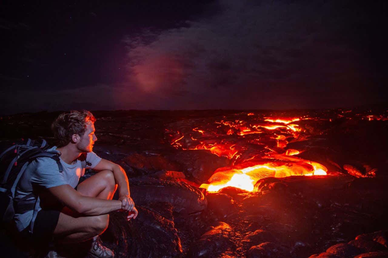 A male tourist wearing a backpack in a kneeling position, looking at bright lava on the ground beside him, with its light  creating on orange hue on his face.