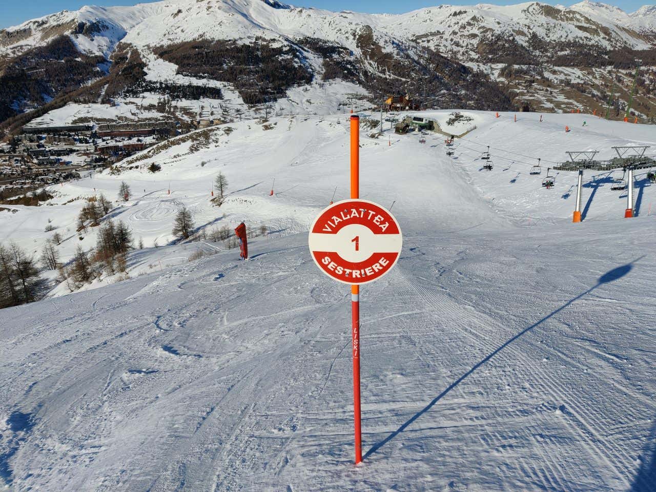 Un palo rosso con un cartello tondo che indica che ci troviamo a Sestriere, sullo sfondo una pista da sci in montagna