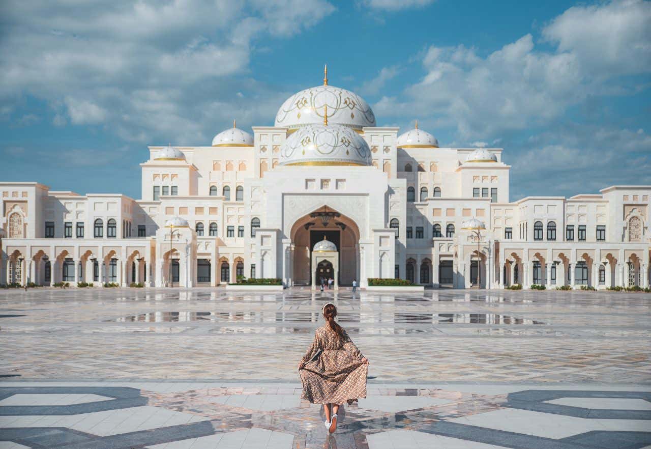 Una donna di spalle nel centro di una vasta piazza dai pavimenti decorati con disegni geometrici, e sul fondo un grande palazzo in pietra bianca e decorazioni dorate