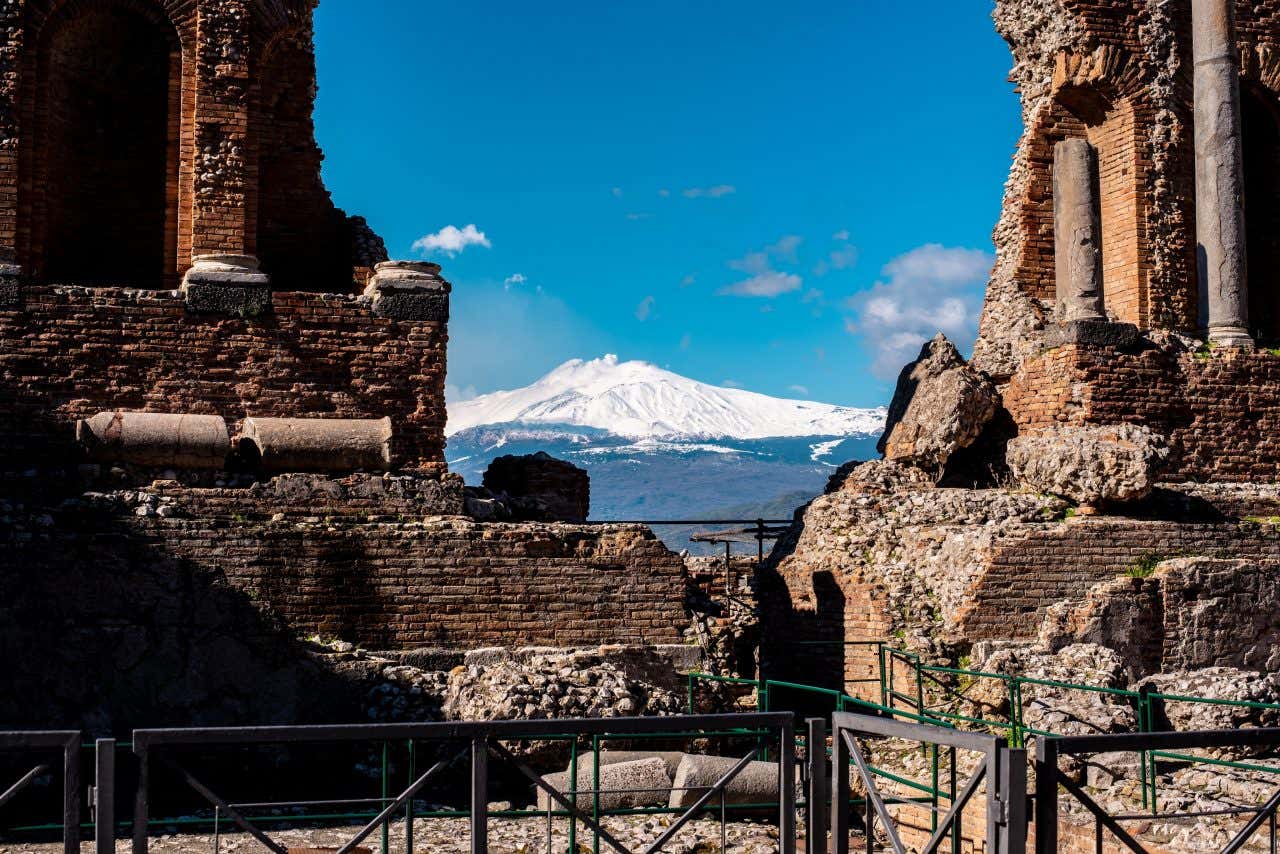 In primo piano i resti di un antico tempio romano, mentre sullo sfondo si vede il vulcano Etna completamente coperto di neve