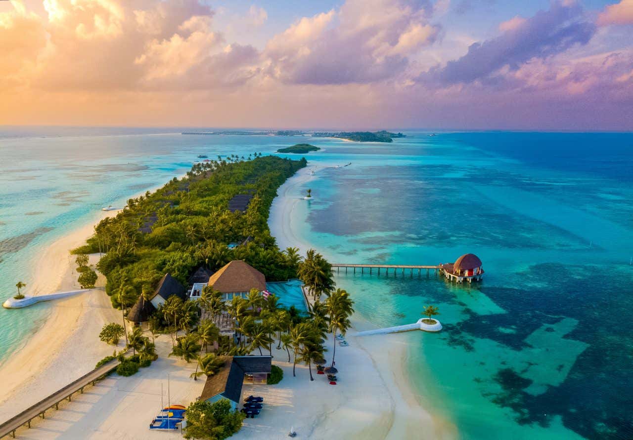 An islandin the Maldives as seen from the air, with clear blue waters on either side and a cloudy orange and pink sky in the background.