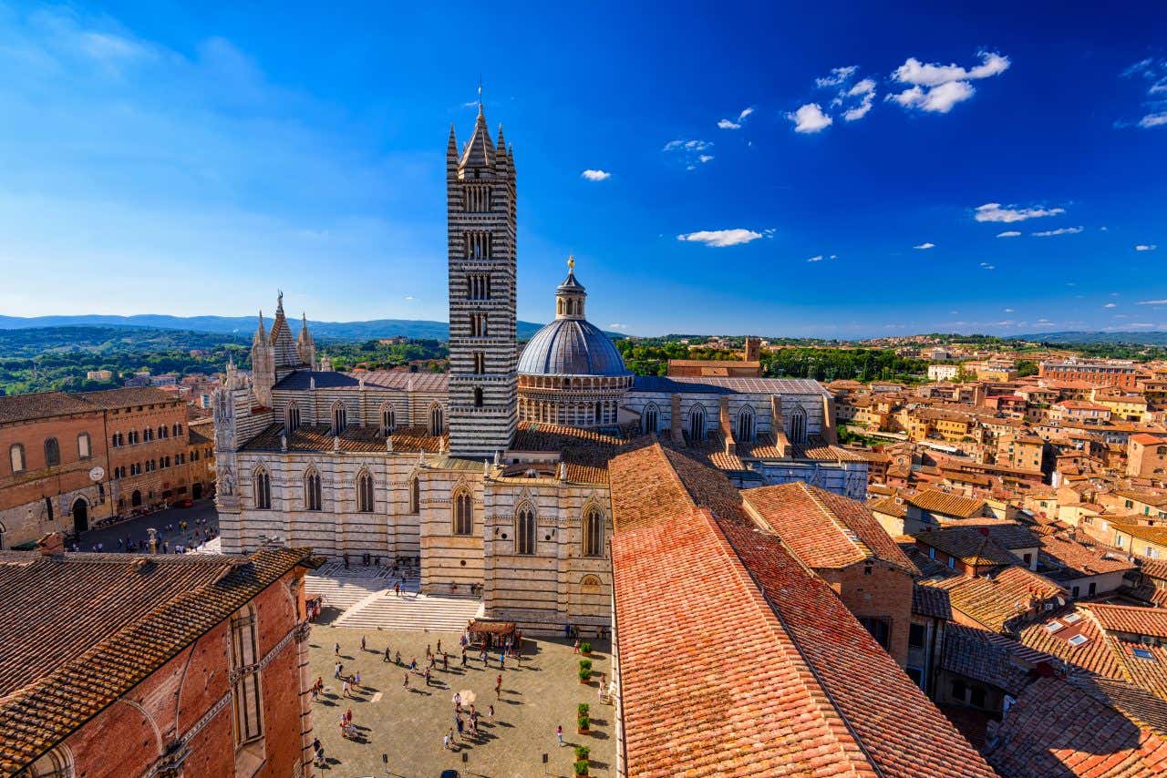 Una grossa cattedrale affacciata su una piazza, con cupola e un campanile, finestre gotiche e decorazioni che alternalo strisce di marmo bianco a strisce di marmo nero