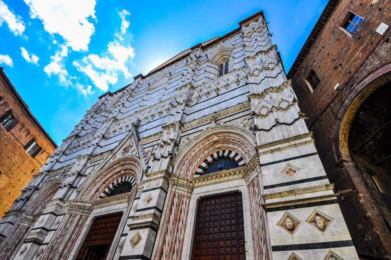 La facciata del Battistero di Siena ripresa dal basso in un giorno di sole, è bianca con sottili strisce nere, archi decorati in stile romanico e finestre gotiche