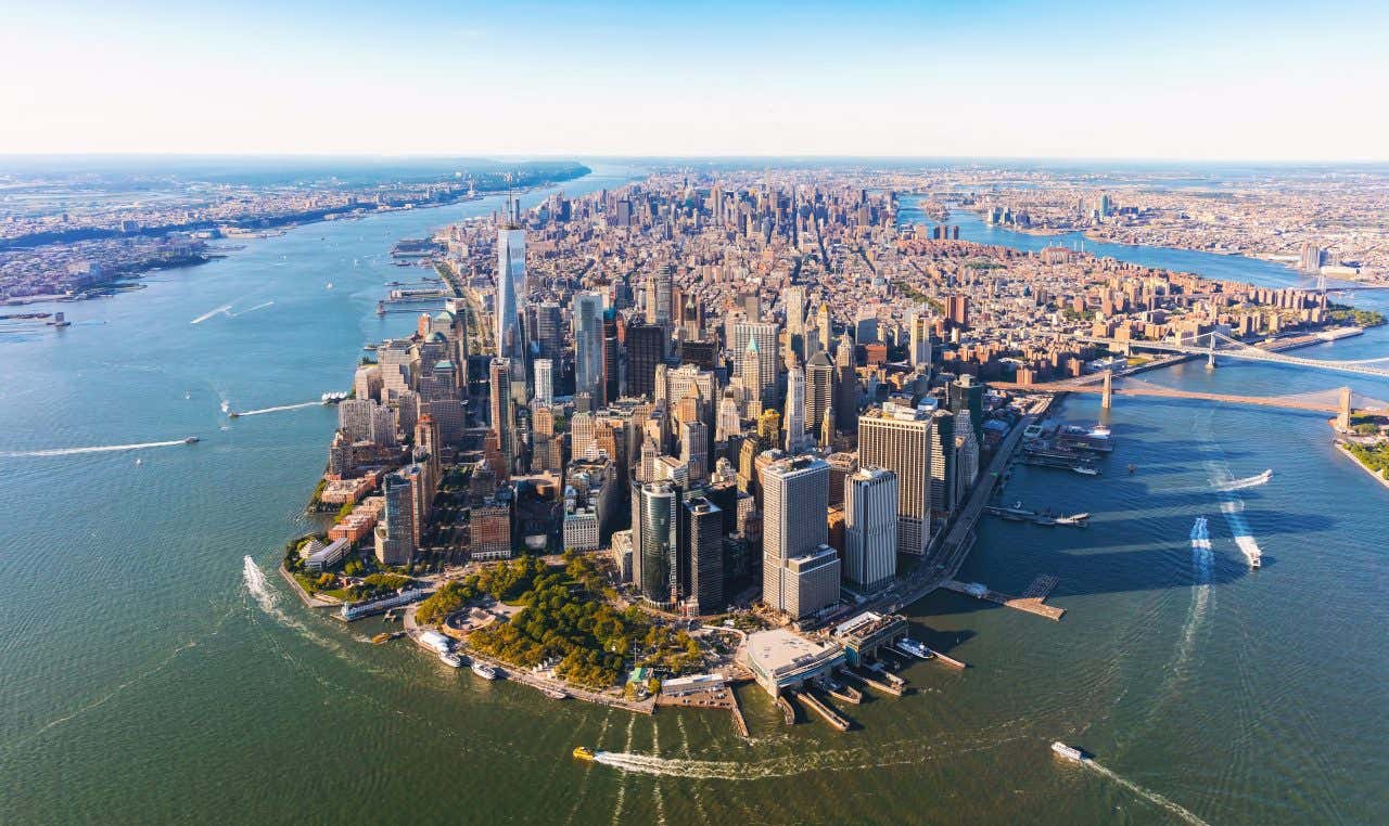 New York City as seen from above, with the river wrapping around Manhatten island and boats in view.