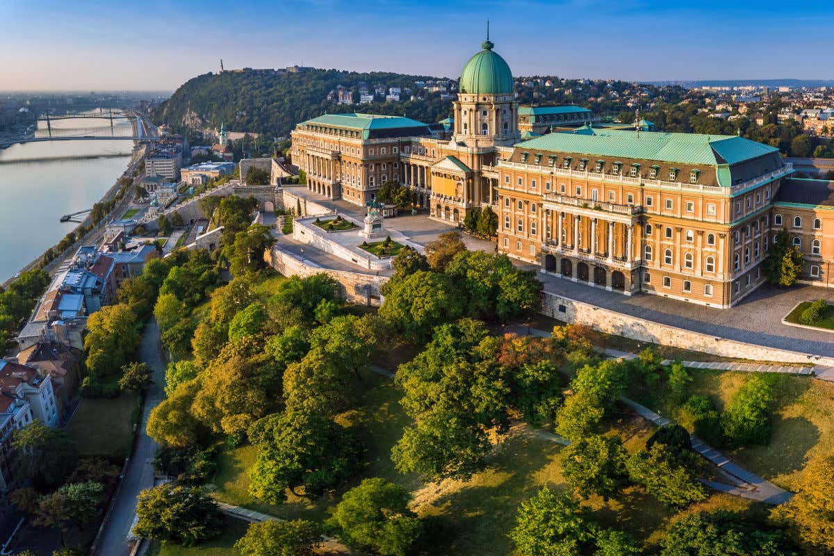 Vista aérea del castillo de Buda, con varios edificios y una gran cúpula rodeada de colinas y árboles frente al río Danubio