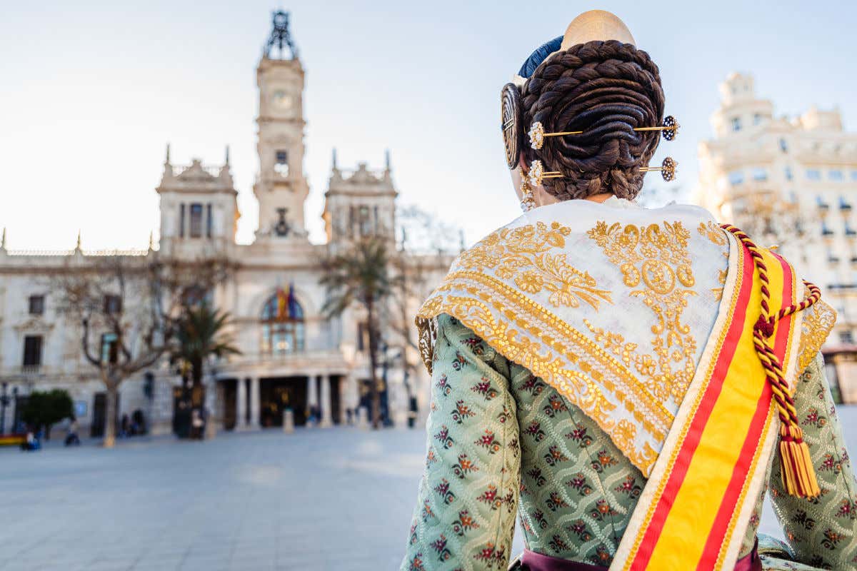 Una mujer de espaldas a la cámara vestida con el traje tradicional de fallera frente a la fachada del Ayuntamiento de Valencia