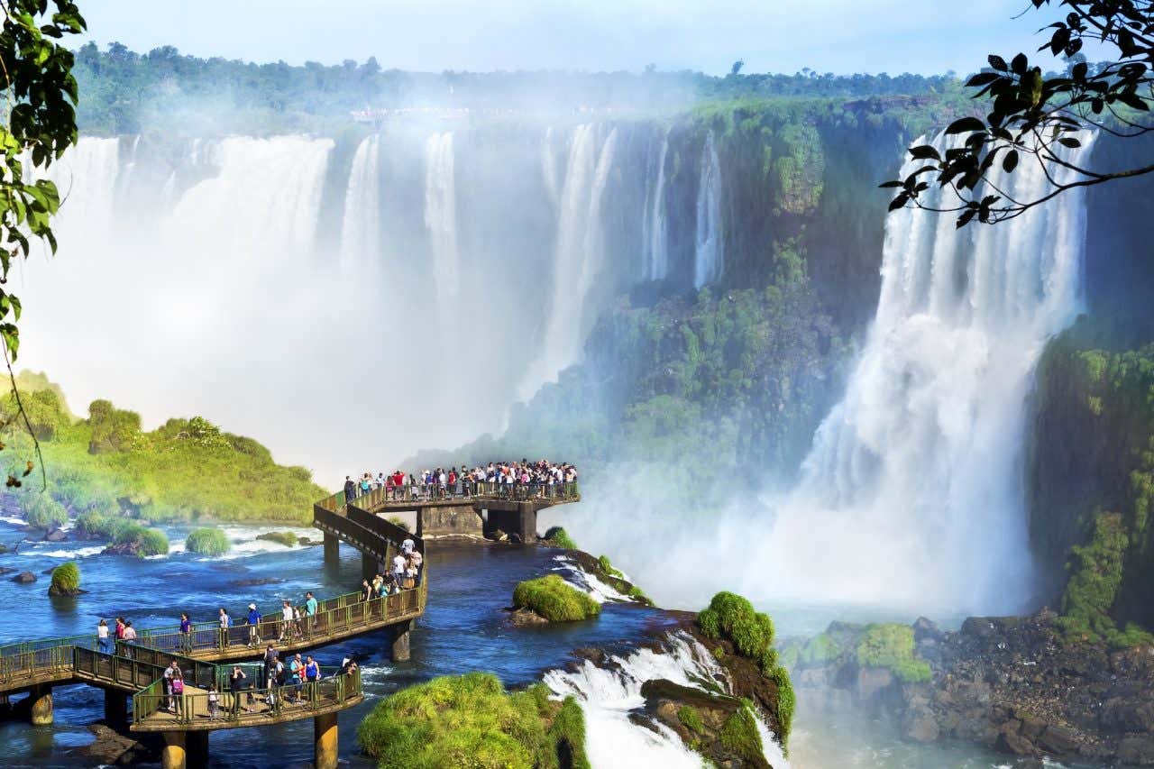 Turistas passeando por uma passarela sobre as águas com as Cataratas do Iguaçu ao fundo